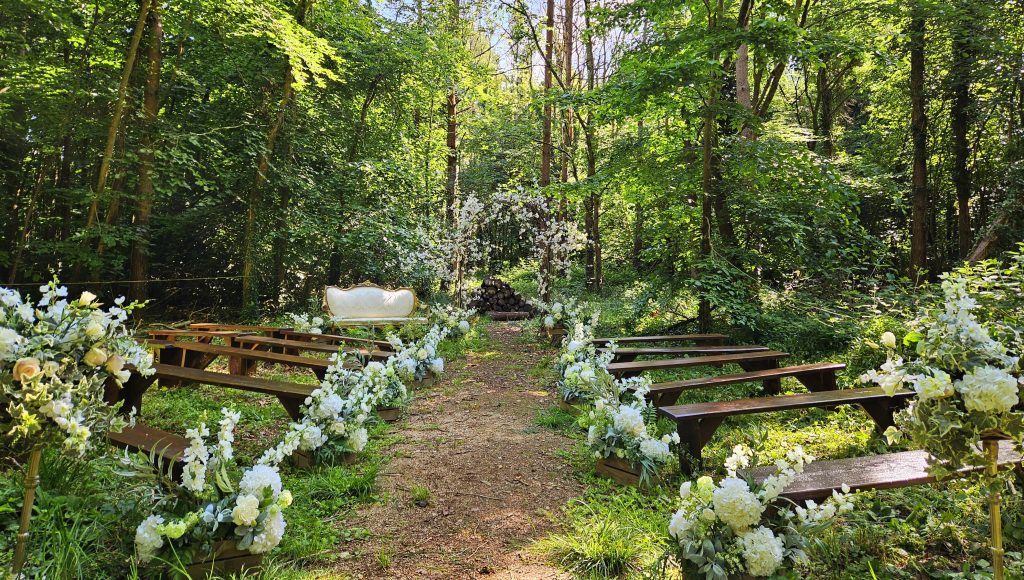 Wild woodland wedding aisle with rustic wooden benches and rustic wooden crates filled with luxury white and green faux fake flowers lining the aisle. At the end of the aisle directly in front of the aisle is a wicker and blossom branch arch and to the left sits our gorgeous white and gold vintage sofa all available to hire