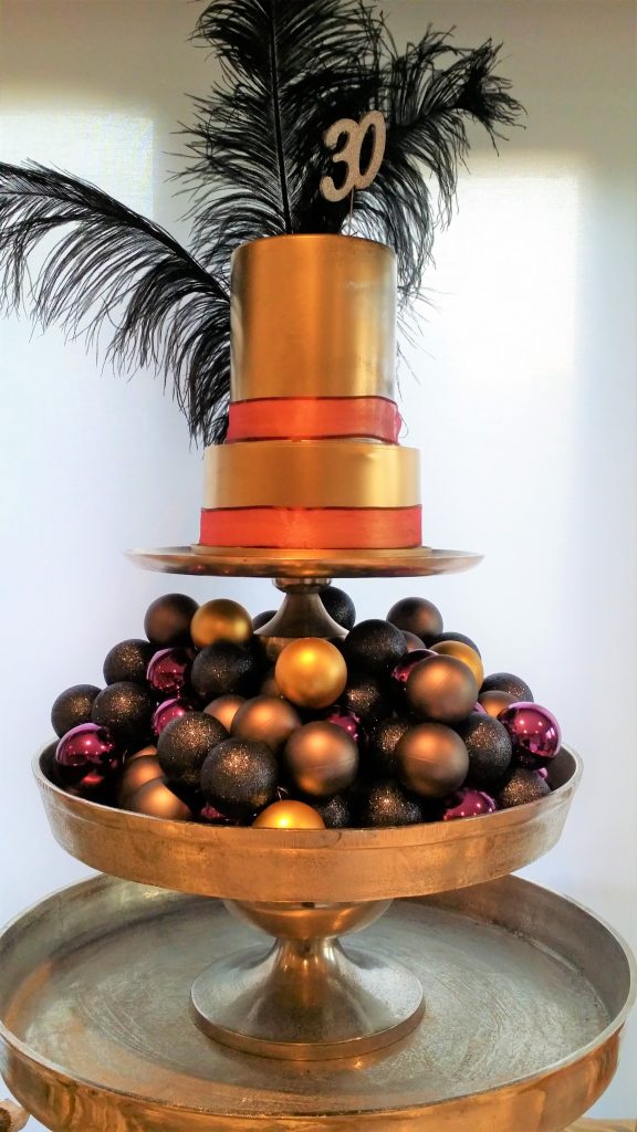 a grand layered three tiered silver cake stand. The middle is filled with purple, black and silver baubles, the top layer is a gold frosted two tier cake with a red ribbon around the bottom of each layer and large black ostrich feathers coming out of the top and a silver glittery 30 candle all available to hire
